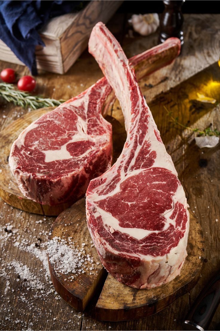 Two thick bone-in tomahawk ribeyes on a wooden board with salt and rosemary